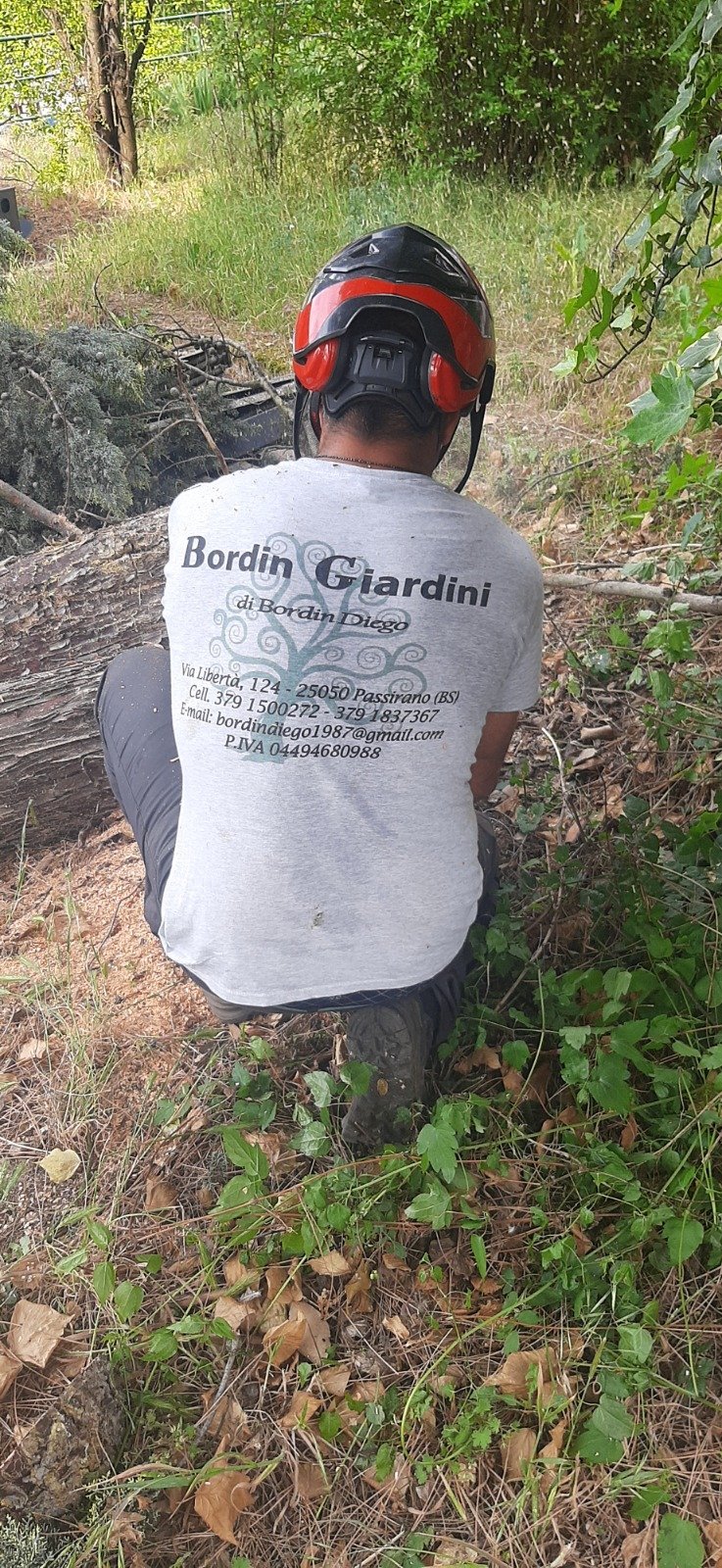 Person wearing red and black helmet and white Bordin Gardini t-shirt standing on forest trail surrounded by green vegetation