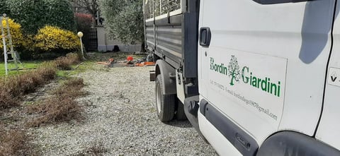 White Giardini work truck parked on a gravel driveway beside landscaped garden beds with shrubs and yellow flowers